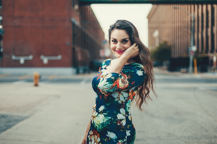 Miriam is wearing a colourful, flowery dress and standing in the middle of an urban center. She is putting her hair behind her ear and smiling at the camera.